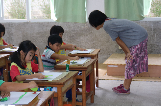 教室內，一位女性老師彎身指導坐在木製課桌前的小學生。多名學生正在桌前進行書寫或繪畫活動，窗戶旁有綠色布幔，牆上貼有教學掛圖，教室呈現活躍的教學場景。