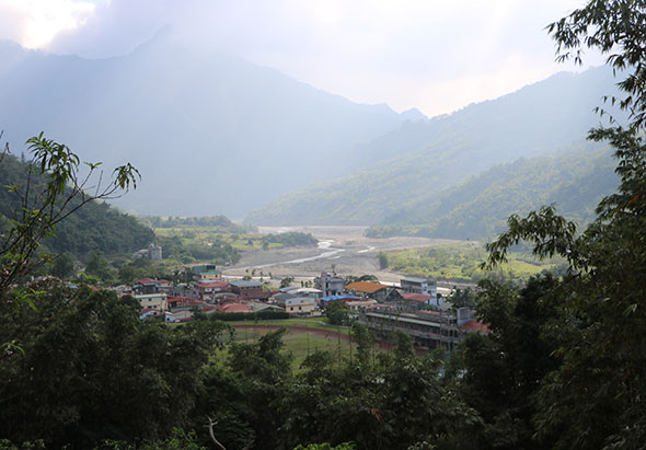 山谷地帶的村落全景。前景為茂密的綠色植被，中景為紅磚房屋、建築群和蜿蜒的河流，背景為層層疊疊的青山。天色晴朗，山水秀麗，呈現寧靜的鄉村風光。