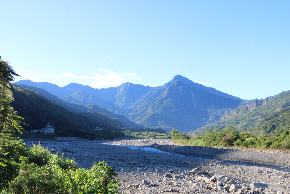 壯闊的高山峽谷風景，清澈溪流蜿蜒流過河床，兩側高聳的藍灰色山峰聳立天際，山坡覆蓋翠綠植被，遠處可見建築物，晴朗天空映照著寧靜的自然美景。