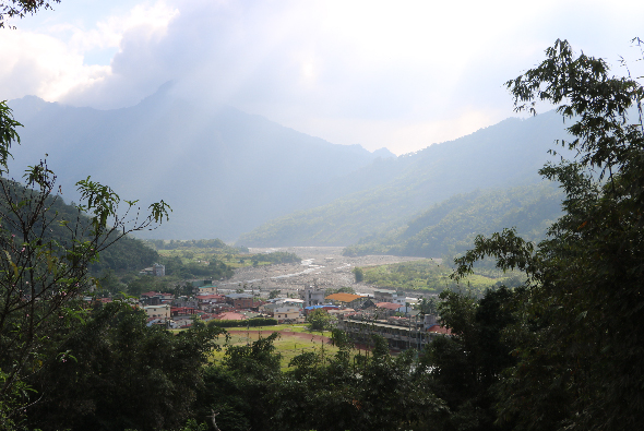 山谷景觀，遠景為層疊的青山，山腰籠罩薄霧，山下為河流蜿蜒流經的村落，房屋密集分佈，綠色田地點綴其間，天空多雲。
