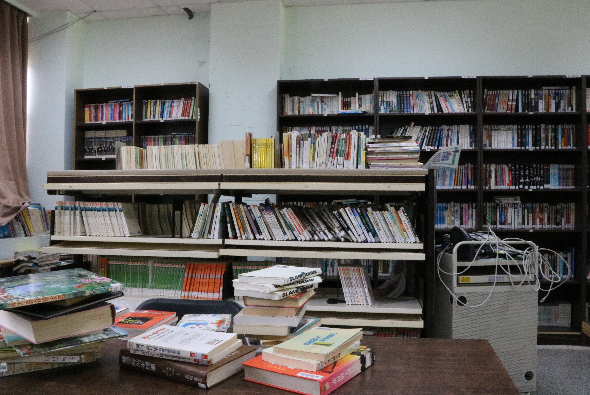 圖書館或書籍收藏室，牆上有多個黑色書櫃放滿各類書籍，中間有淺色層板架，前方木桌堆放著多疊書籍和雜誌，右側放有垃圾桶，展示豐富的閱讀資源與學習環境。