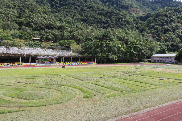 山區運動場景。前景為紅色跑道和綠色草坪，草坪上有白色螺旋線條紋。背景是看台建築和藍色休息室，遠處為茂密森林和山丘。場地設施完整，適合田徑和球類運動。