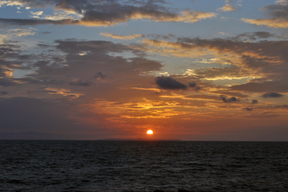夕陽西下於平靜的海面上，太陽呈現金黃色圓盤，逐漸沉入海平線。天空被橙紅與藍灰色雲彩渲染，光線在雲層間投射出絢麗色彩，營造出寧靜壯麗的傍晚海景氛圍。