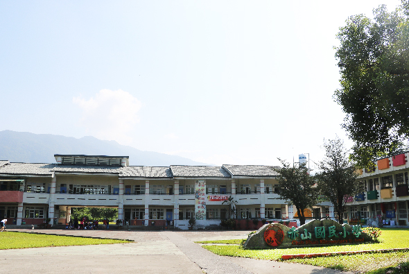 開闊的廣場中央，一座藍白色現代建築物坐落於中間，兩側為紅色商店招牌。前方草坪上有綠色裝飾花壇，背景可見遠山。晴朗天氣，整體呈現小鎮商業廣場的景象。