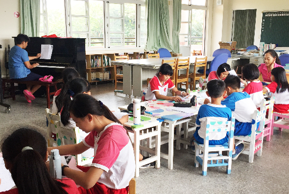 教室內，多名穿著紅白制服的小學生坐在白色小桌椅前進行美術創作。左側有學生在鋼琴前練習，背景是寬敞的窗戶和綠色窗簾，呈現活潑的教學環境。