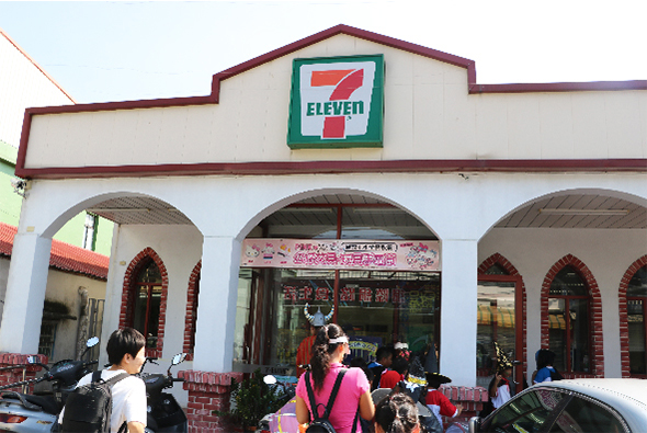 7-Eleven便利店外觀，白色建築搭配紅色磚砌拱形入口，頂部懸掛綠色7-Eleven標誌。店門前有數位顧客駐足，停放多輛機車。