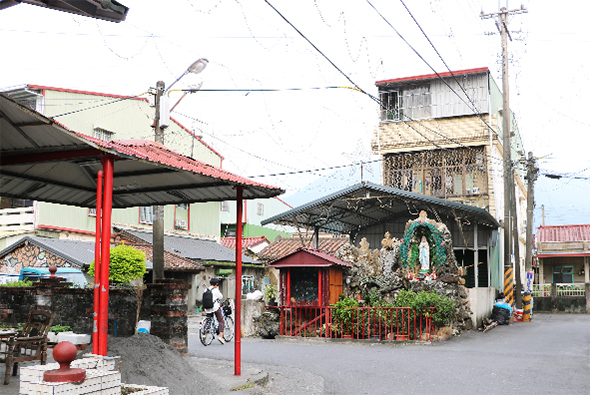 台灣鄉村街景，左側有紅色柱子的停車棚，中央紅色門窗建築物，右側為老舊水泥樓房，電線縱橫交錯，道路兩側植有綠色植物，呈現傳統農村聚落風貌。