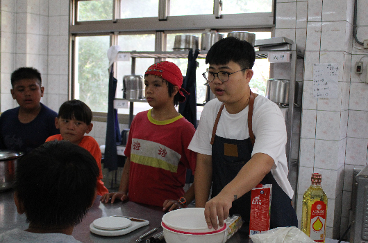 廚房裡，一位穿著黑色圍裙、戴眼鏡的年輕男性正在準備食物。他身旁站著穿著紅色衣服戴紅帽的少年，以及其他幾位參與者。桌上擺放著白色餐盤、鍋具和調味料，窗戶背景可見室外景色。這是一場烹飪教學或美食活動。