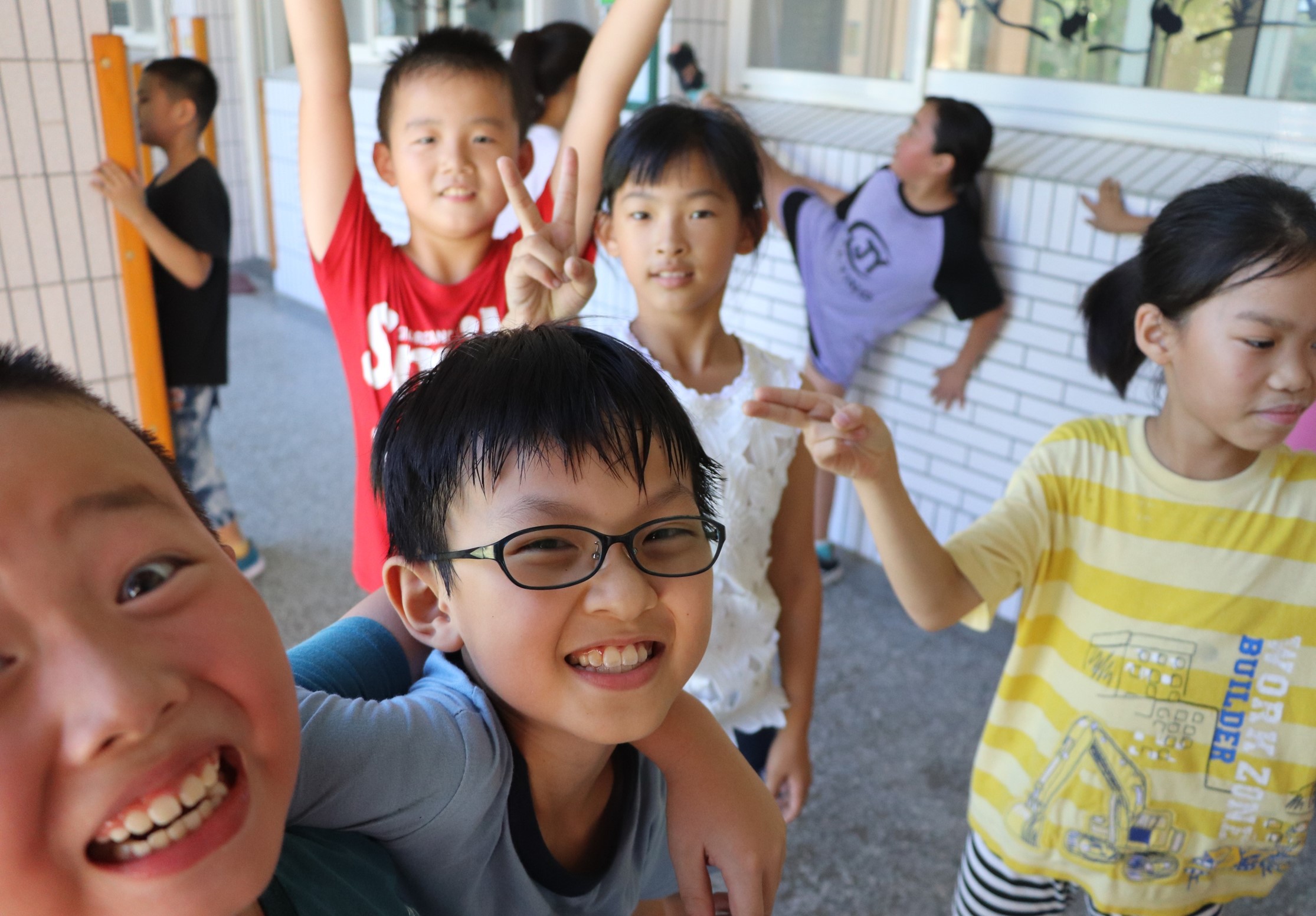 一群小學生在室內走廊聚集，穿著紅、黃、藍等彩色衣服，面帶笑容，有人舉手做出勝利手勢，營造歡樂活潑的校園氛圍。