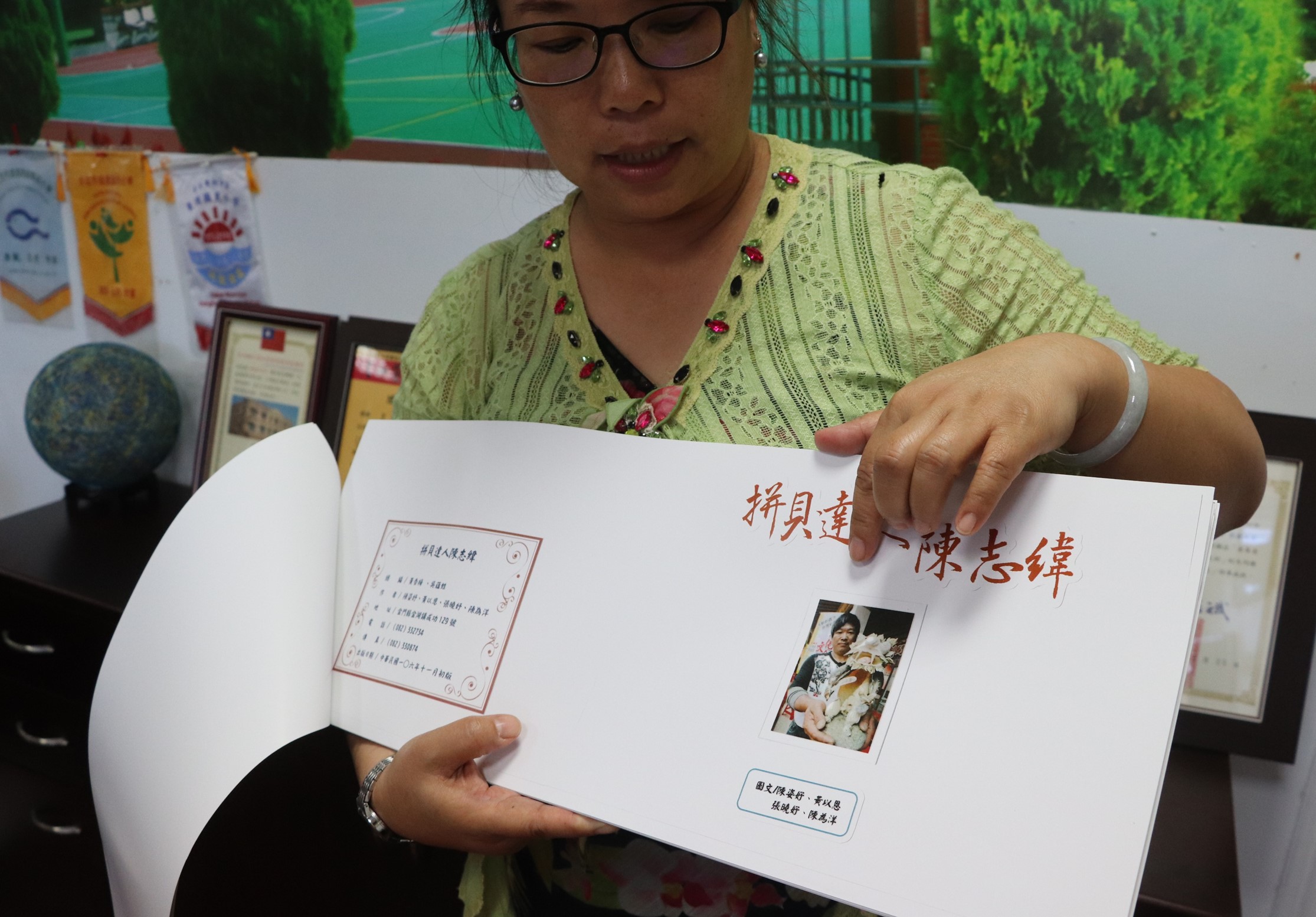 一名戴眼鏡的女性在室內展示一本開啟的紀念冊，封面印有紅色中文標題「摯愛選一陳志綸」，內頁貼有小照片和文字說明。背景可見獎狀、地球儀等物品。