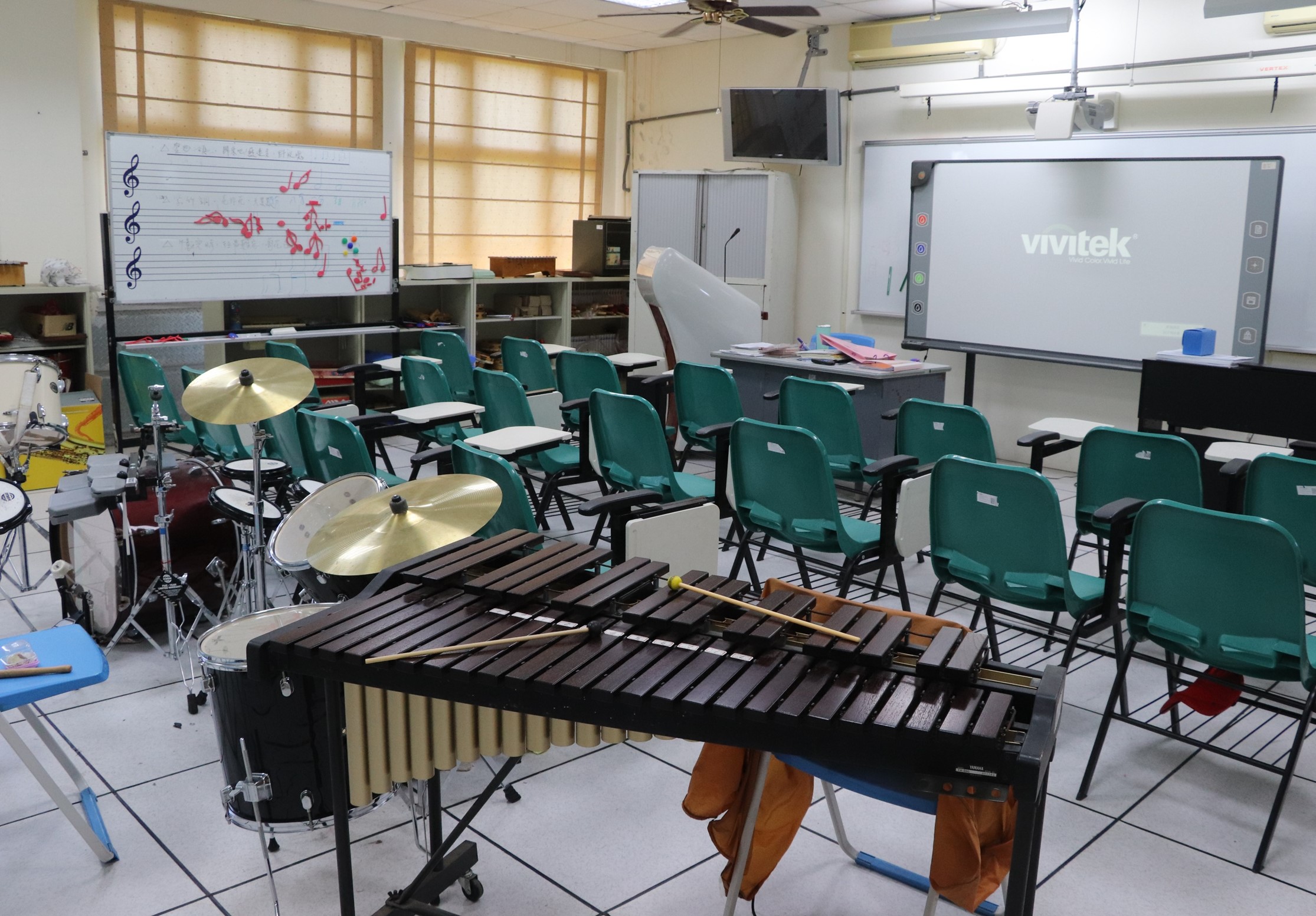 音樂教室內擺放著木琴、鼓等樂器，綠色折疊椅排成半圓形供學生使用。牆上有五線譜板和電子白板，教室設有投影機和風扇，適合音樂課程教學。