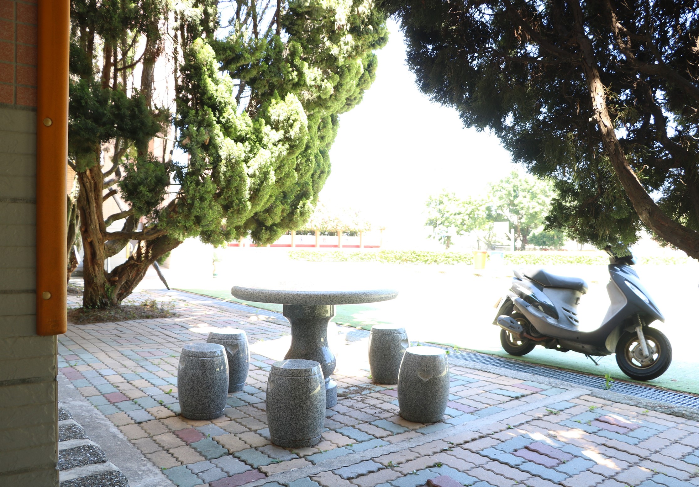戶外休閒區域，石製圓桌周圍環繞著六個圓形石凳，供人們坐著休息。濃密的樹木提供遮蔭，地面鋪著紅棕色磚石。右側停放著一台白色機車，背景可見紅色欄杆和空曠的綠色草地。