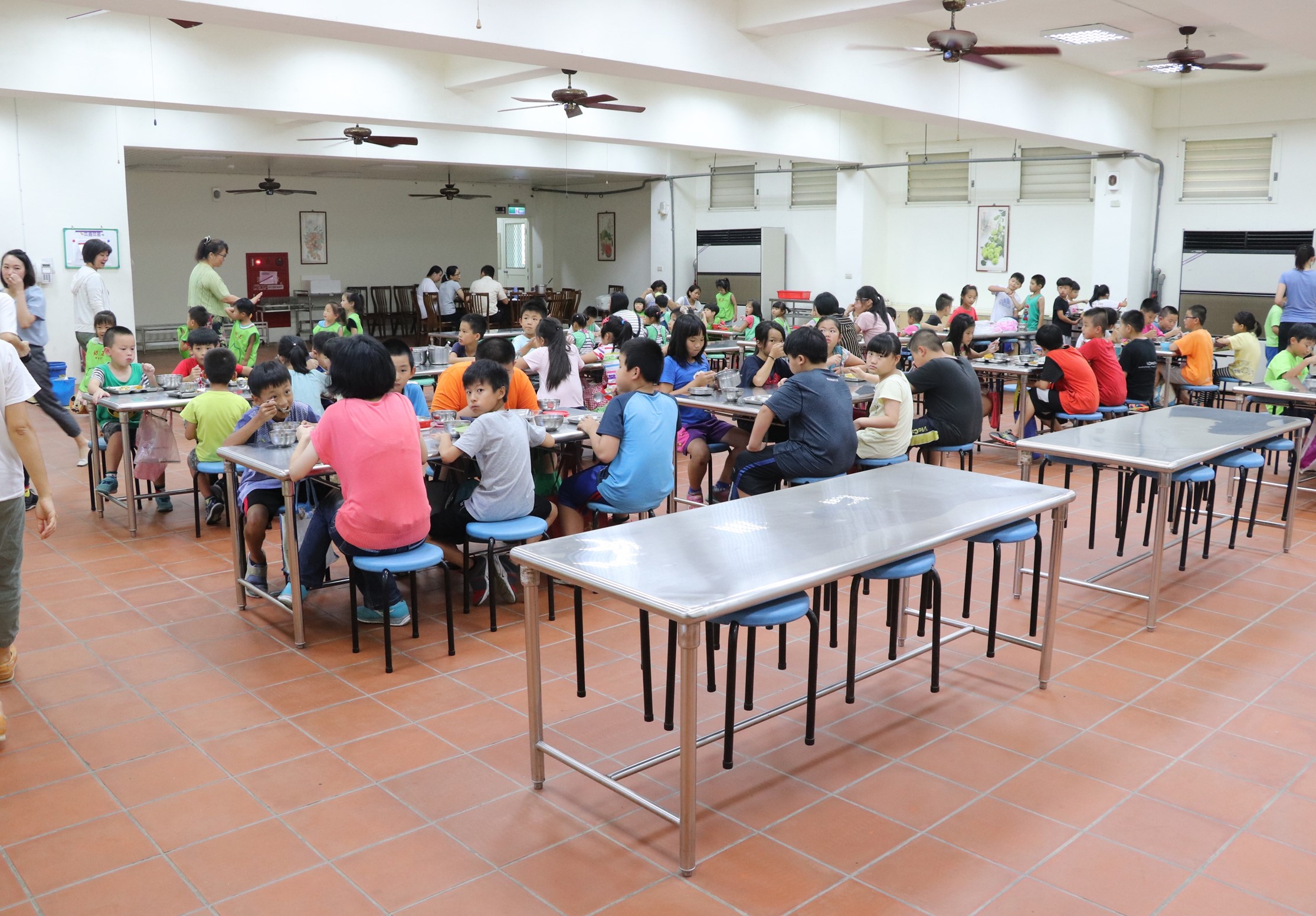 學校食堂內，大約五十多位穿著彩色衣服的小學生坐在多張長桌前用餐。場景寬敞明亮，天花板有多台風扇。成人在一旁監督和協助。整體呈現溫馨的集體用餐情景。