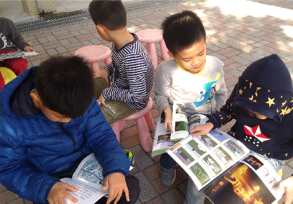 四名兒童在戶外磚製地面上圍坐成一圈，低頭查看手中的彩色雜誌或宣傳冊。兒童穿著藍色、黑白條紋和深藍色外套，專注地翻閱印刷物。背景為磚製人行道和建築物。