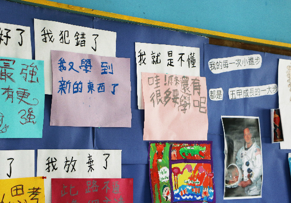 教室布告欄展示學生的中文書法和繪畫作品，貼有多張寫滿中文字句的紙張，包括粉紅色、白色和紅色背景的作品，以及色彩繽紛的兒童畫作和學生照片。