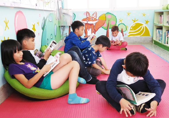 兒童教室內，六名小學生坐在粉紅色墊子上進行閱讀活動。牆上有彩色動物壁畫和黃星星裝飾。綠色豆袋椅供學生使用。書架擺放在教室角落。孩子們手持書籍專注閱讀。