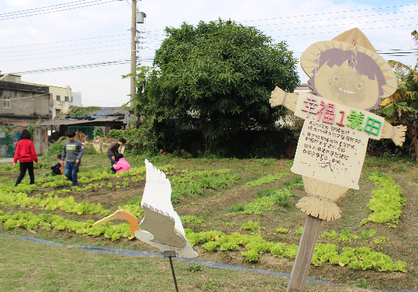 農村社區菜園活動現場，數位穿著彩色衣服的人在種植蔬菜。田間豎立著木製稻草人和標示牌，背後有大樹和建築物。田地種滿綠色蔬菜苗，展現農耕教育的情景。