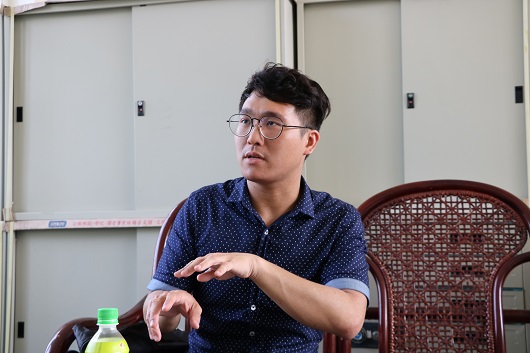 一名戴著眼鏡、穿著深藍色點點襯衫的男性在室內演講或分享。他坐在木製椅子上，面向左側，身旁放著綠色寶特瓶。背景為白色推拉門和講台，環境顯示為教室或會議室。