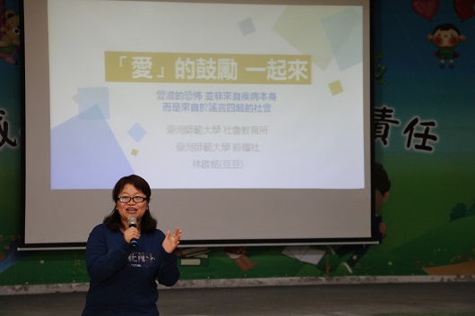 一位戴眼鏡的女性在教室講台前進行演講，身後的投影幕上顯示標題「愛的說關一起來」，幕後展示教育相關內容。講者右手持麥克風，左手做出手勢，與觀眾互動。教室牆面貼有中文字面。