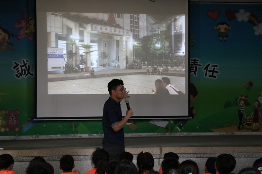 一位男性講者站在教室前方，手持麥克風進行演講。身後大螢幕播放一部影片，顯示一條繁華的街道景象。下方坐著一群觀眾，認真聆聽。教室牆上貼有中文字和裝飾品。