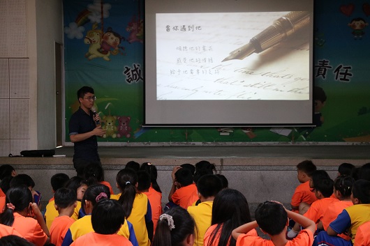 教室內，身穿黑衣的男性講師站在投影幕前進行教學，幕上顯示書法相關的中文內容和毛筆圖片。坐在前排穿著橙色和黑色制服的學生約二十人，認真聆聽講師的課程講解。