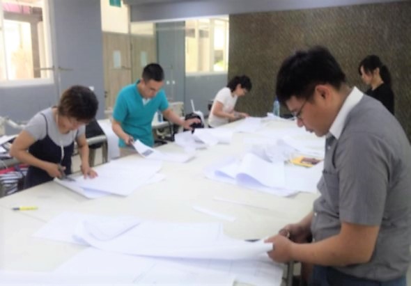一群人圍在大白色工作台旁，正在進行手工藝或設計活動。前景一位穿著灰色上衣的男性低頭專注地操作手中的材料，左側女性也在認真工作，背景還有其他參與者和工作人員。室內光線充足，牆壁貼有紋理壁紙。