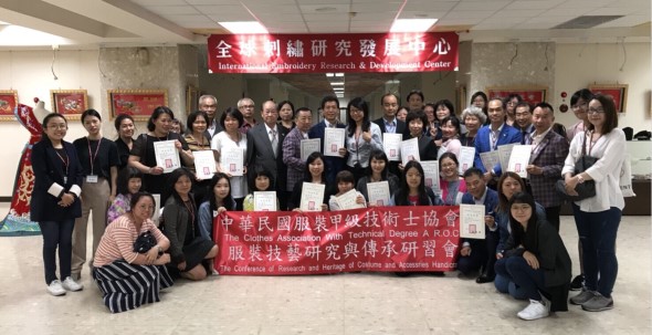 國際刺繡研究發展中心舉辦活動，約五十位參與者於室內合照。眾人手持證書或文件，中央擺放寫有中文文字的紅色布條。背景牆上掛有紅色標誌牌匾，天花板為格子狀。參與者穿著正式服裝，展現活動圓滿成功。