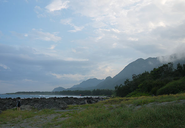 海邊景色，前景為岩石灘和草地，遠方山脈在雲霧中若隱若現，海水在左側，天空佈滿白雲，一人站在岩石灘上眺望遠方，呈現寧靜的自然風光。
