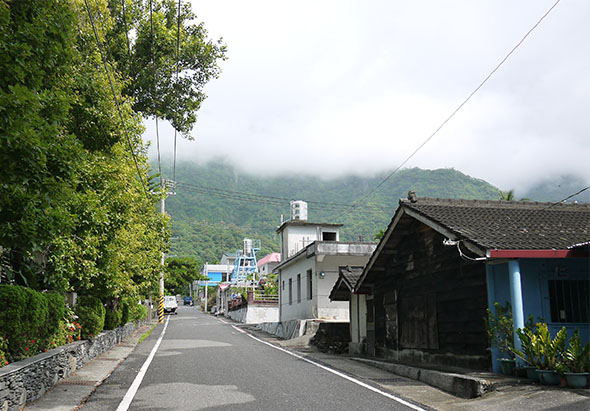 山區鄉村道路景象，兩側有傳統建築和現代房屋，左側種滿綠色植物，遠方山峰被雲霧籠罩，天色陰沈，電線杆立於路旁，呈現寧靜的鄉間風貌。