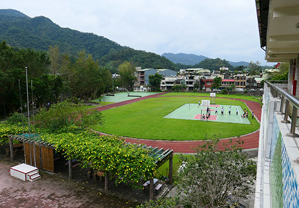 俯視學校運動場景。紅色跑道環繞綠色草坪，中央白色區域有學生進行體育活動。右側建築物、左側綠化區和背景山景可見。晴天環境，設施完善。