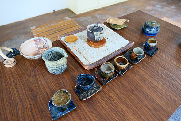 木質桌面上展示的茶具和陶瓷藝品。左側有白色盤子和淺藍色陶罐，中央是白色方形托盤配黑色蓋碗，右側排列多個深藍色和棕色的小陶罐和杯具，背景可見木製工作台和白色牆面。