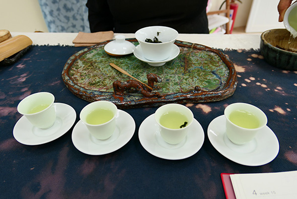 茶藝展示會現場，深藍色布景桌上擺放四杯裝有淡綠色茶湯的白瓷杯盤，中央為裝有茶葉的古樸茶盤和茶荷，背景可見茶具和參與者。