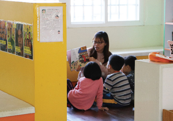 幼兒教室內，一位女性教師坐在地上，向三位穿著粉紅色和條紋衣服的幼童展示繪本故事書。黃色隔板和綠色牆壁構成溫暖的教學環境，窗戶採光良好。