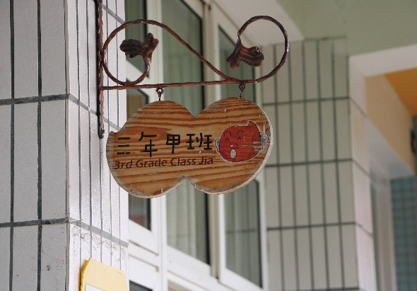教室門上懸掛的木製班級牌，呈雙圓形設計，上面刻有「三年甲班」和「3rd Grade Class Jia」的英文字樣，搭配紅色圓形裝飾，牌子由鐵製彎曲裝飾架支撐，背景為白色百葉窗和綠色牆面。