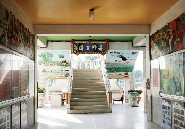 寺廟或宗教建築的內部走廊，中央為寬敞的階梯通往上層。天花板為黃色，兩側牆壁貼有宗教海報和告示牌。正上方懸掛紅底金字的中文牌匾。階梯兩旁放置植物盆栽和宣傳資料架。