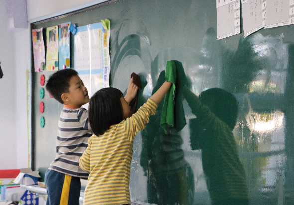 兩名小學生在綠色黑板前，穿著條紋衣服的學生用綠色抹布擦拭黑板。教室環境中可見布告欄和教學用具。