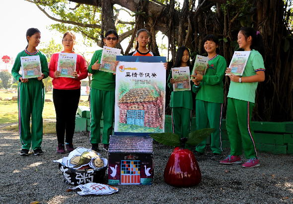 六位穿著綠色運動服的學生在大樹下展示繪畫作品和宣傳海報。前方地上擺放著裝飾品和紅色物件，背景為草坪和樹木。此為校園或社區環保宣傳活動的合照。