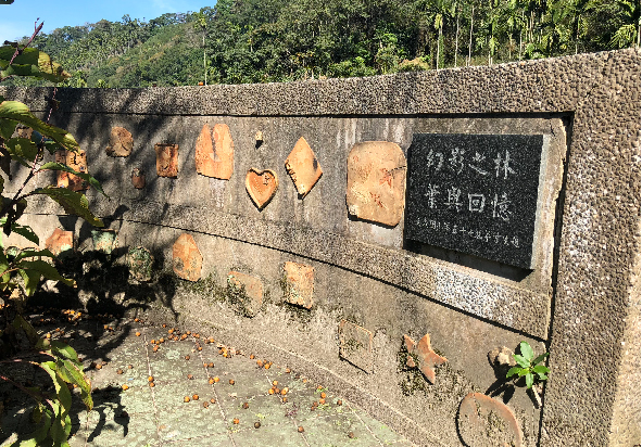 石製紀念碑，上方刻有心形和其他形狀的浮雕，右側嵌有黑色紀念牌匾，前方放置鮮花，背景為綠樹覆蓋的山坡，碑座周圍有小植物生長。