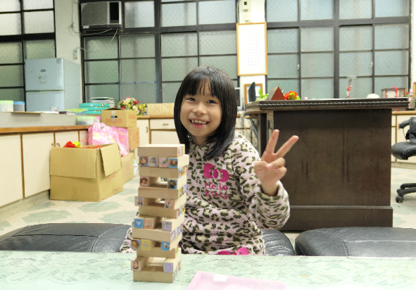 一名穿著豹紋上衣的小女孩在教室內做出勝利手勢，面前堆疊著木製積木遊戲。背景可見教室設施、儲物櫃和教學用具，展現兒童遊戲學習的快樂氛圍。