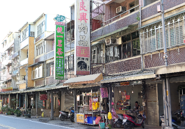 繁華的街道商業區，多層樓房林立，各式招牌和標誌密集排列。街道底層為商店鋪面，有捲簾門、招牌和商品展示。樓上為住宅，窗戶和防盜窗密布。街道停放機車和人力車，呈現典型的亞洲都市街景。