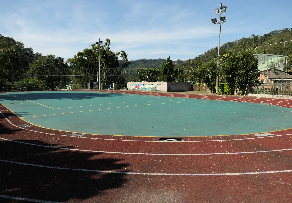 室外多功能運動場地，綠色塑膠跑道中央為藍綠色手球場，周圍有紅色跑道。場邊有照明燈桿、觀眾席和建築物。背景是茂密的樹林和山丘，天空晴朗。