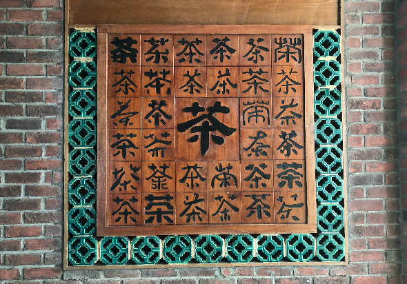 磚牆上貼著木製方形裝飾板，中央為7×7網格排列，每格刻有繁體「茶」字或其變體，中心最大字體為「茶」。四周邊框飾以綠色幾何花紋瓷磚，整體構成茶文化主題藝術裝置。