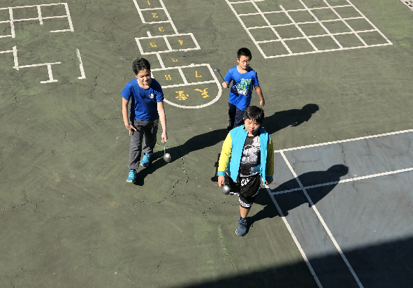 四名學童在操場上進行戶外活動。三人穿著藍色運動服，一人穿著彩色外套，站在白線標記的遊戲區域內。操場地面繪有數字方格和跳房子遊戲標記，陽光投射出清晰的人物影子。