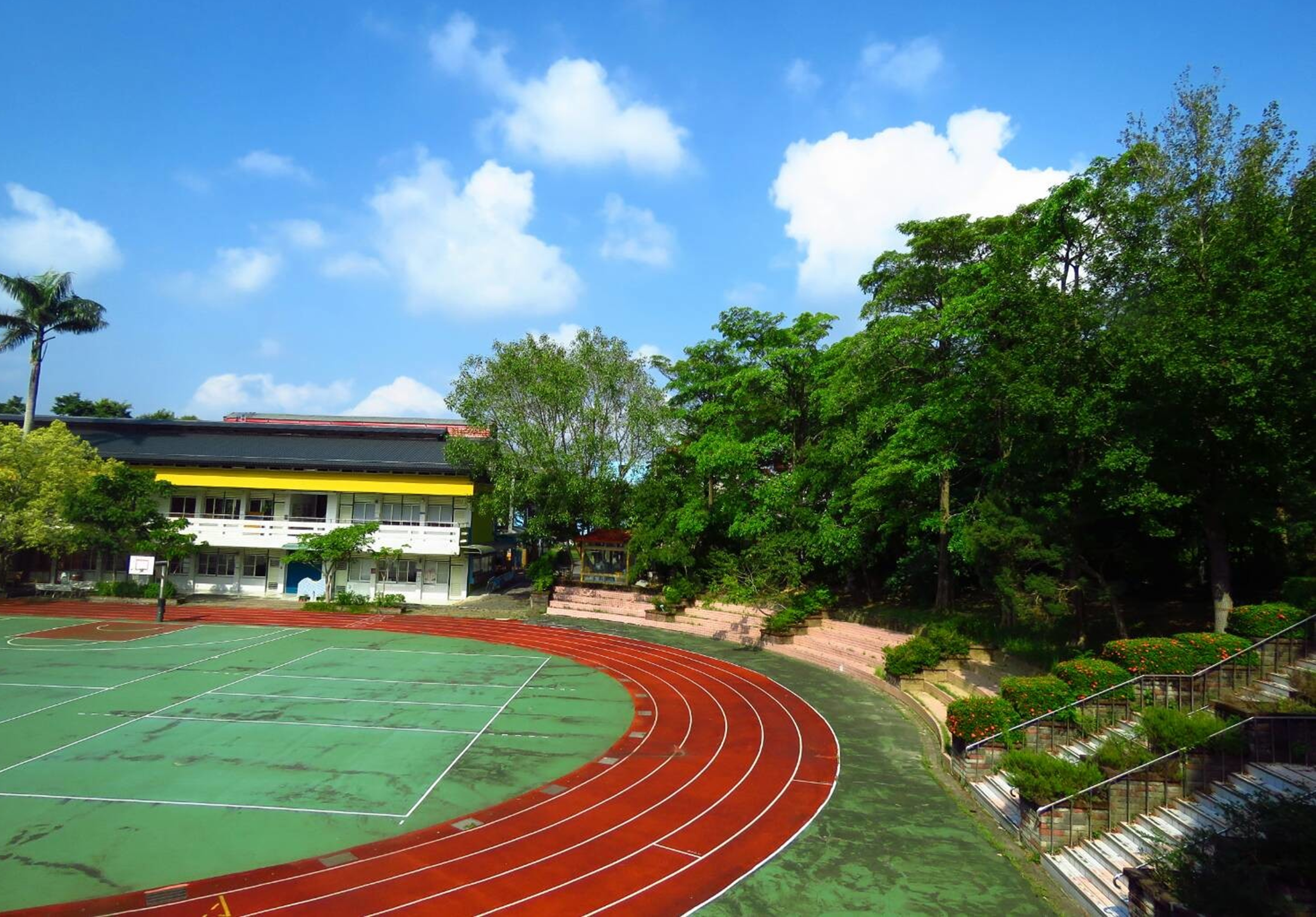學校運動場景，紅色跑道環繞綠色網球場，黃黑相間的校舍坐落於左側，右方為茂密樹林，藍天白雲晴朗，階梯和欄杆通往運動場。