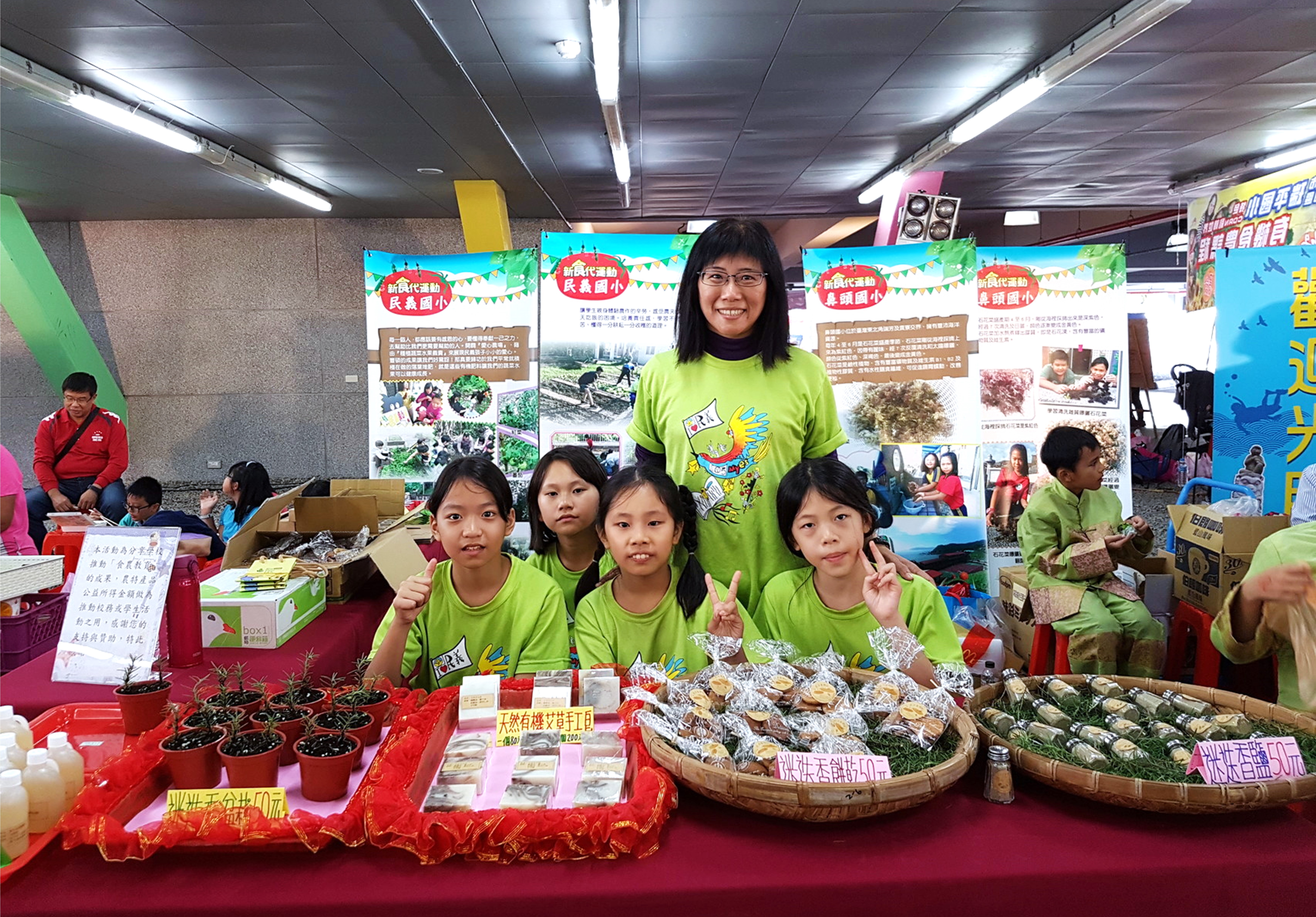室內展覽活動現場，五位穿著綠色制服的女性志工站在紅色展示台前，展示各類農產品和植物盆栽。背景有資訊海報和其他參展攤位，展台上放有竹籃裝的蔬菜、紅色盆栽植物和產品介紹卡片。