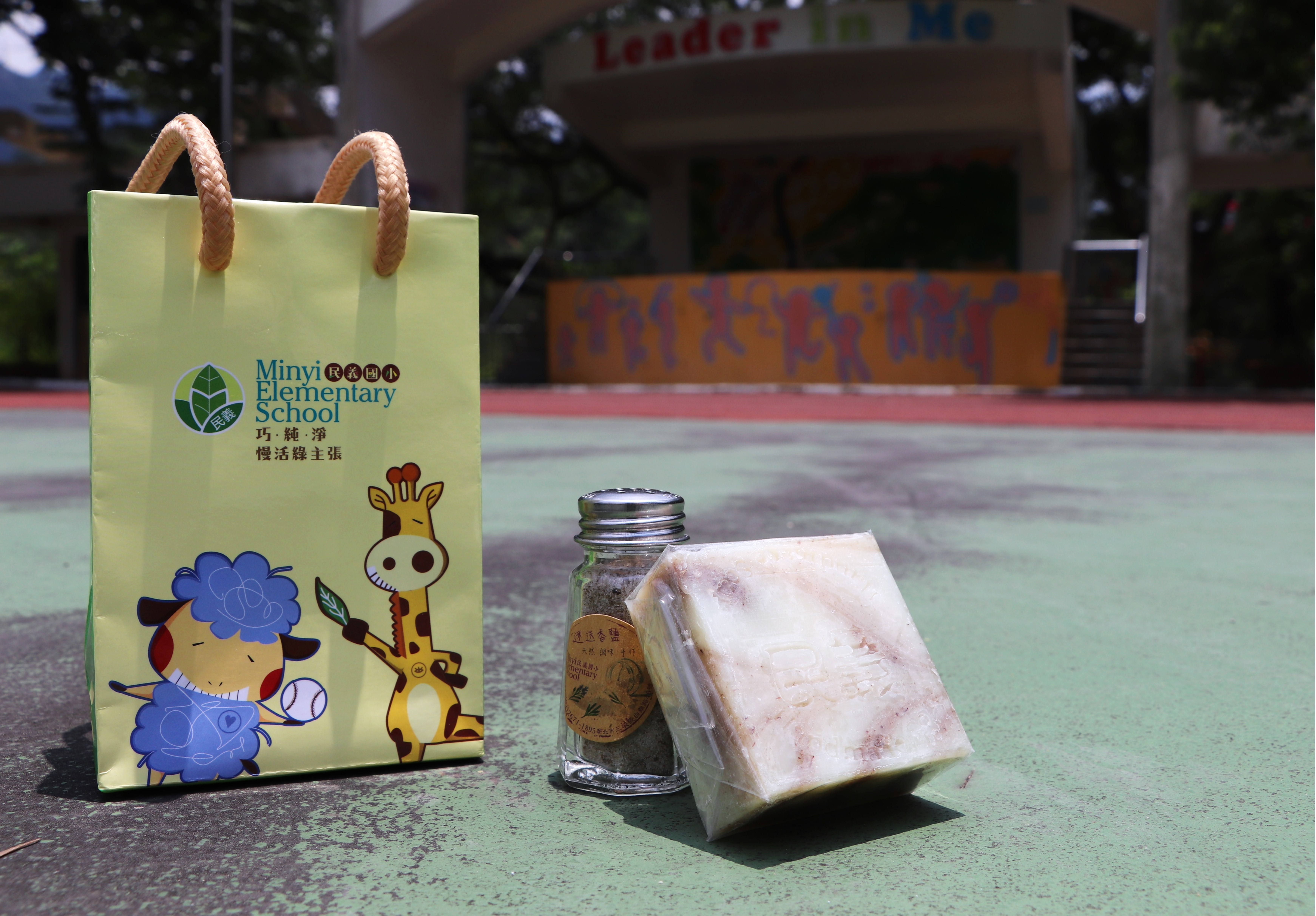 校園廣場上展示民雨小學的黃色紙袋禮品，袋上繪有可愛的綿羊和長頸鹿插圖。紙袋旁放置一塊粉紅色糕點和一瓶玻璃罐裝食品。背景可見校舍建築和紅色操場。