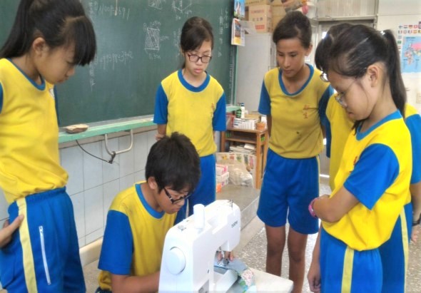 五名穿著黃藍相間校服的學生在教室內圍成一圈，中央男同學正操作一臺白色的科學實驗裝置。背景為綠色黑板和教室佈置，同學們認真觀察實驗進行情況。