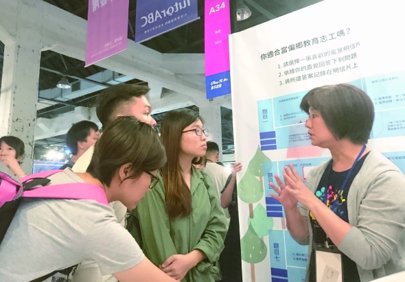 室內展覽會場，一位女性講者向三位年輕女性訪客介紹展示板上的內容。展示板上有中文文字和綠色圖表。背景可見紫色和藍色的展示標牌。現場還有其他參展者在瀏覽展覽。