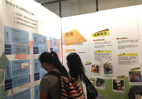 展覽會場中，兩位女性觀看展示板。左側展板介紹工作內容，右側展板以黃色和綠色區域展示「鹿樂」和「偏鄉狀況」主題，附有多張照片和文字說明，呈現教育相關的資訊展示。