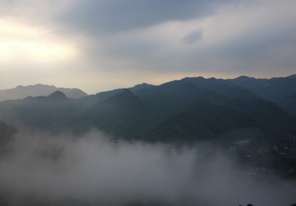 晨霧籠罩的山谷景觀。多層山脈在晨曦中若隱若現，濃霧瀰漫於山谷間，晨光透過雲層灑下，營造出寧靜神秘的氛圍。山脈呈深綠色，遠處漸漸褪色成灰藍色的輪廓。